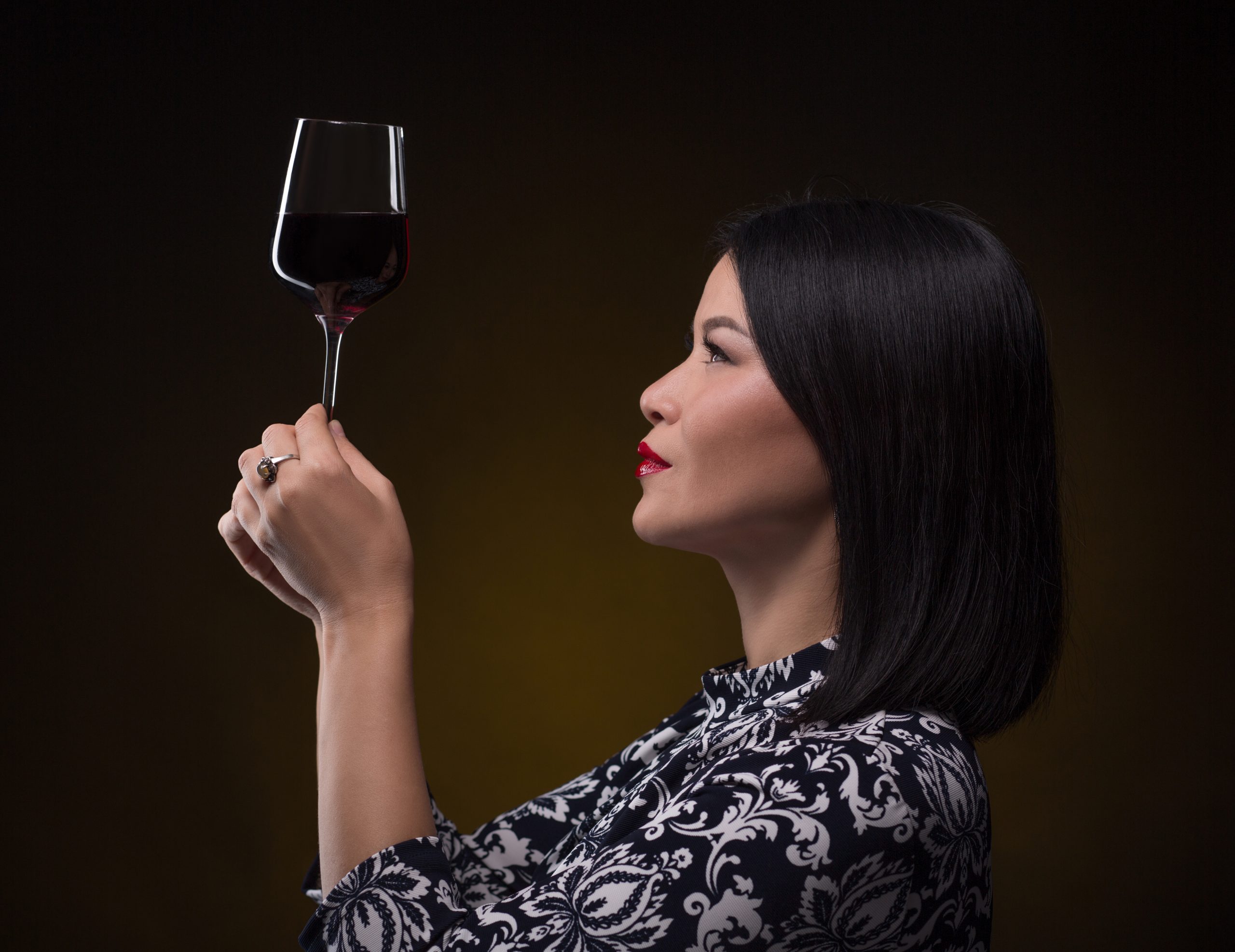 Asian sommelier woman looking at glass of red wine very attentively. Beautiful lady with blach hair holding glass in front of her.